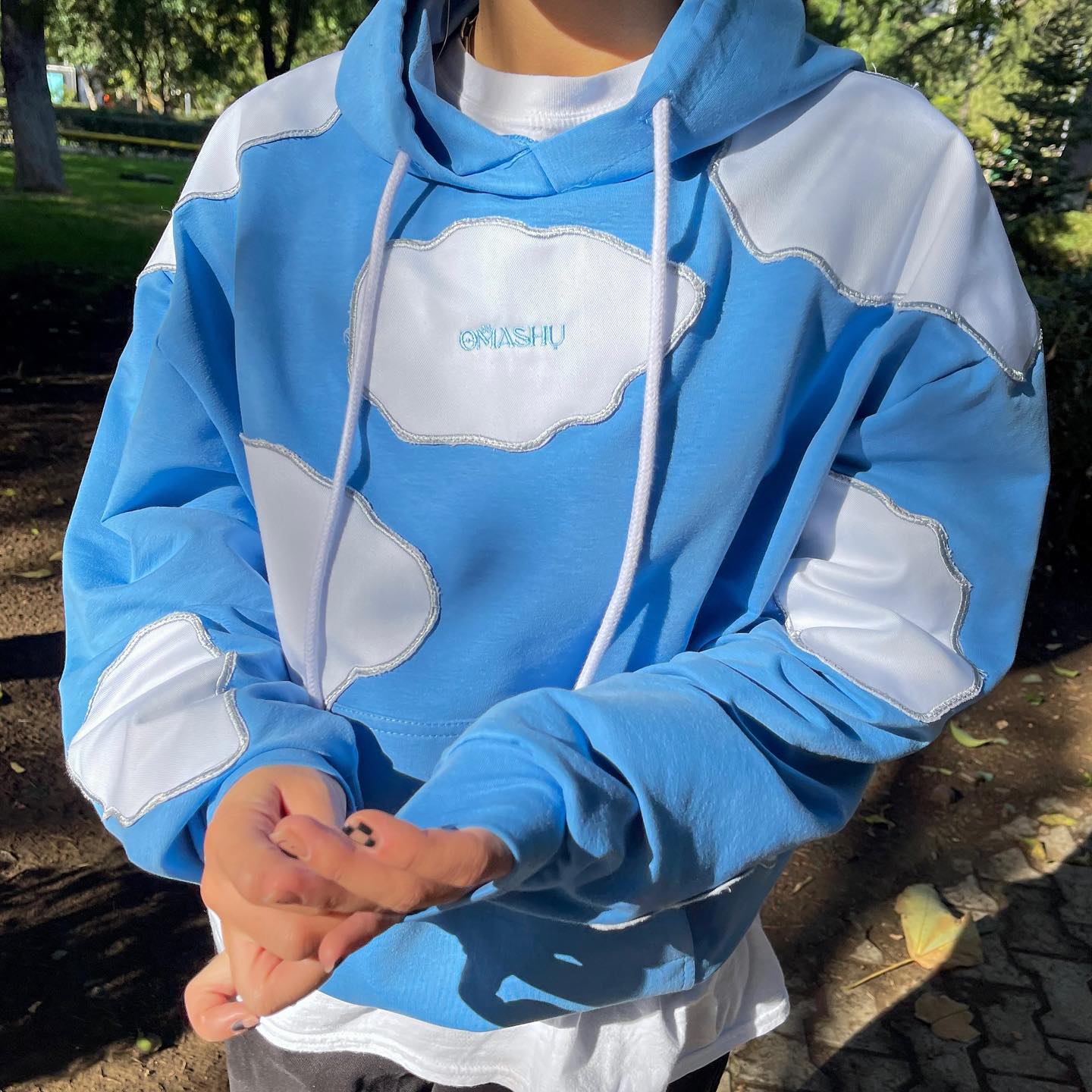 Person wearing blue hoodie with white cloud-like design on front with word ‘OMASHU’ written in sky blue letters and black nails standing in park with trees and grass during the day.