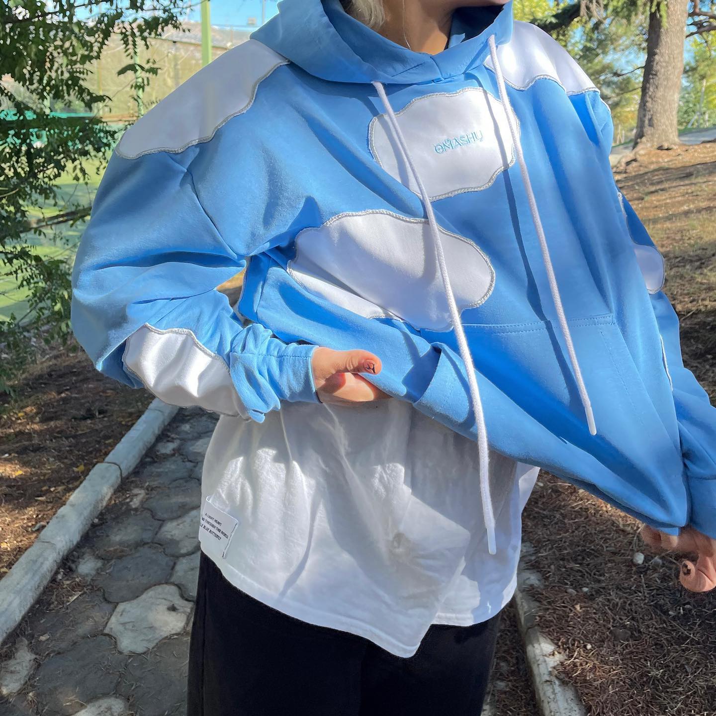 Person wearing blue and white hoodie with white panel on front with ‘OMASHU’ written on it, drawstring hood, and kangaroo pocket standing in park with trees and fence in background during the day.
