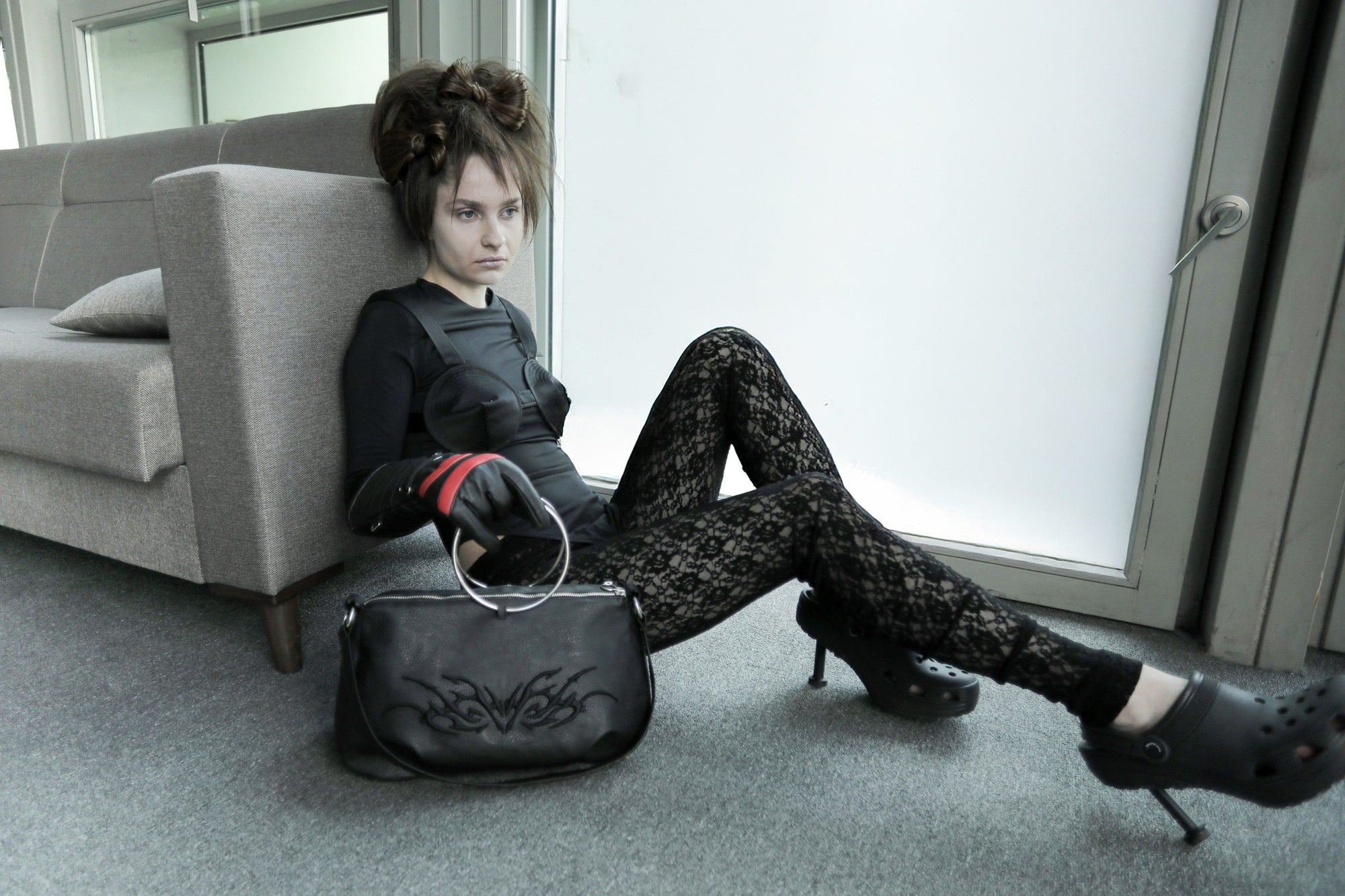 Model in a fashion-forward black outfit crouches, holding the Omashu Anya black faux leather bag with its distinct silver ring handle and embroidered detail, in a modern, window-lit room.