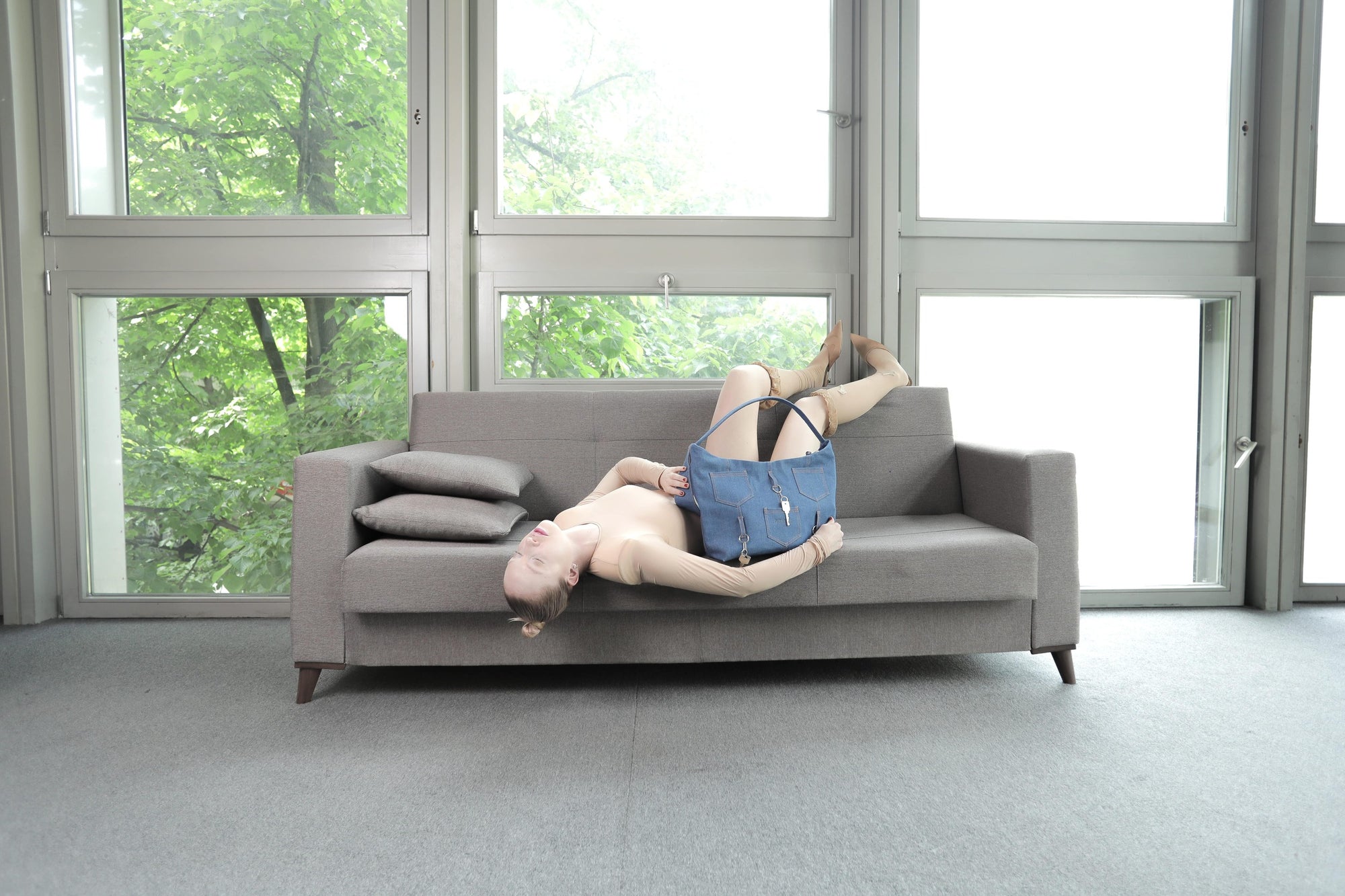 Model lounging on a grey sofa with the Omashu Ines blue denim shoulder bag, showcasing its relaxed style and unique key embellishments in a bright, modern interior.