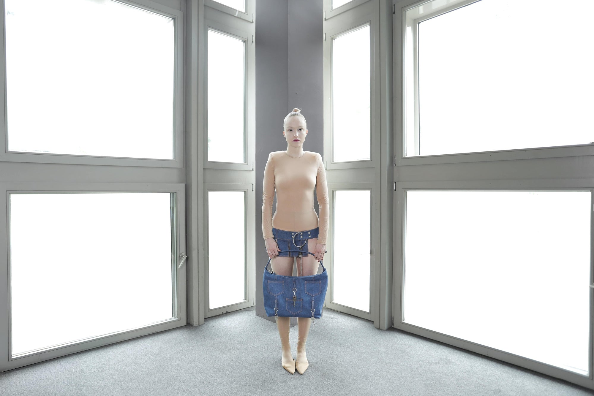 model in a minimalist setting holding the Omashu Ines blue denim bag by its shoulder strap, highlighting its multiple pockets and key accents.