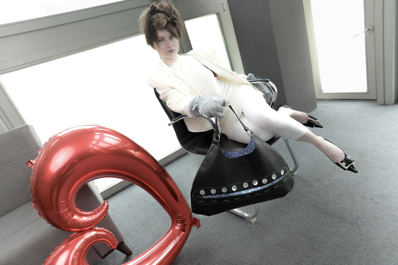 Fashion model lounging stylishly in a chair, the Omashu Sylvie black faux leather bag with its unique button eclipse feature resting on her lap, red heart balloon in background.