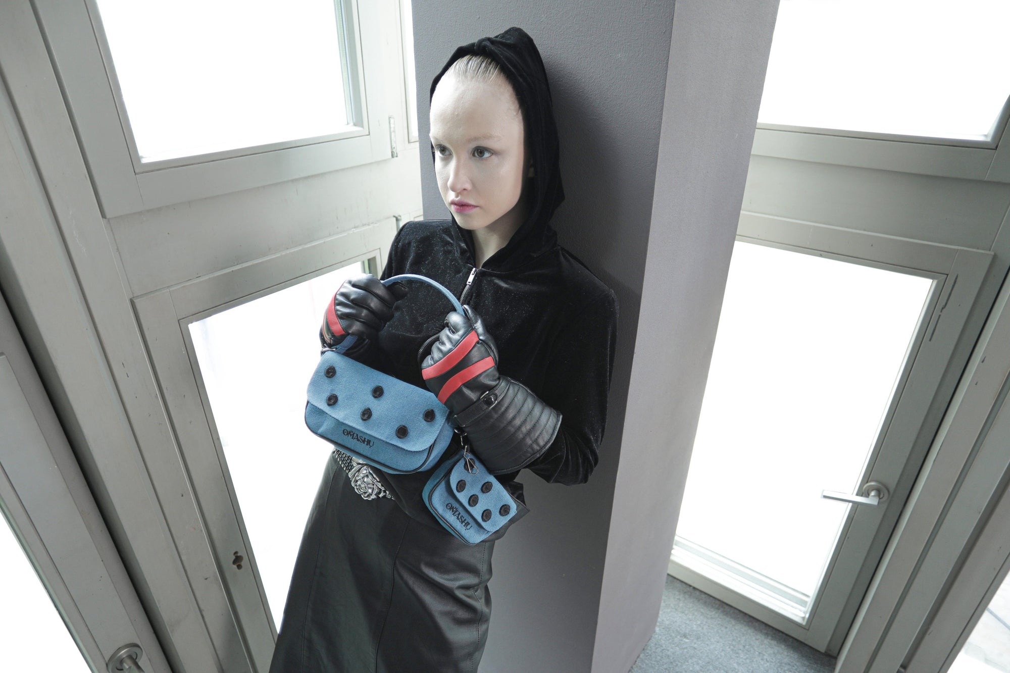 Fashion model in a dark, minimalist outfit showcasing the Omashu Nina blue cotton shoulder bag and attached dot-patterned pouch, held in front.