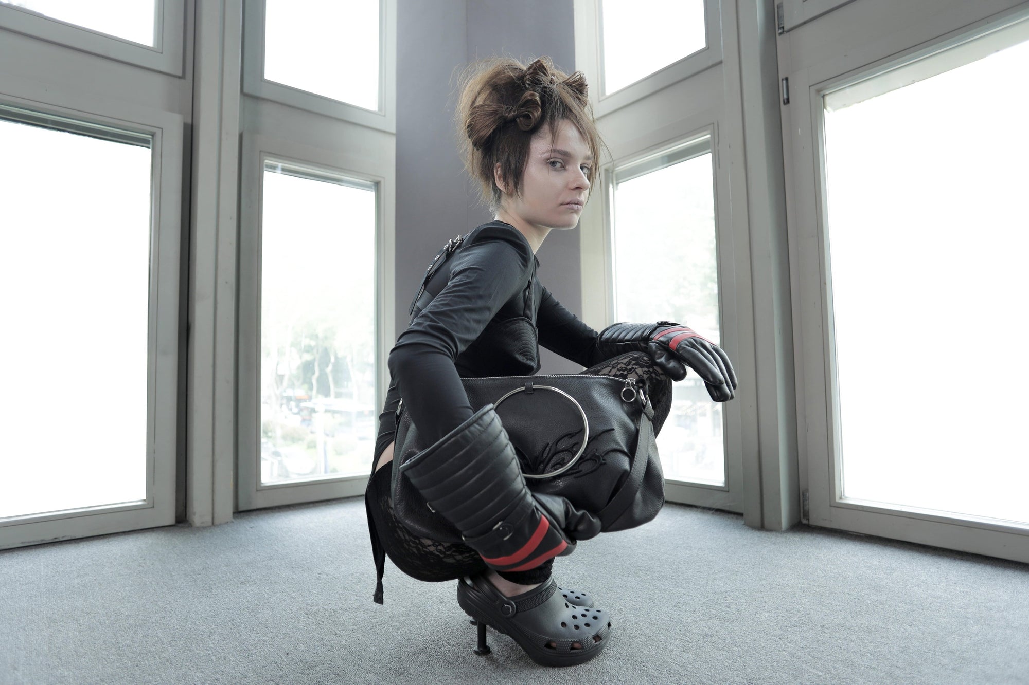Model with edgy styling sits on the floor next to a grey sofa, showcasing the Omashu Anya black ring handle bag with tonal embroidery.