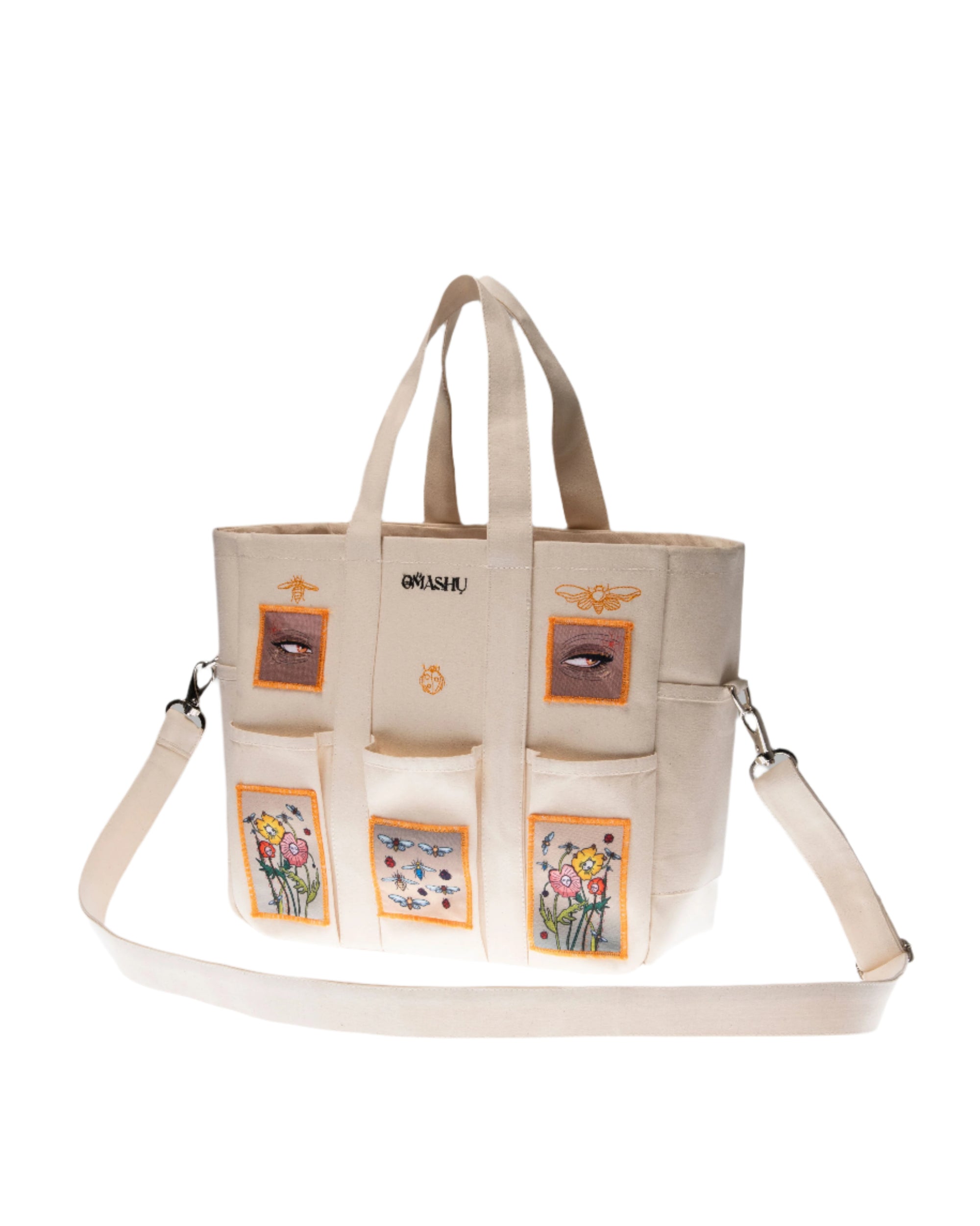 A beige tote bag with multiple pockets, each adorned with different embroidered designs including flowers and a crown, and the word ‘OMASHI’ stitched at the top, against a grey background.