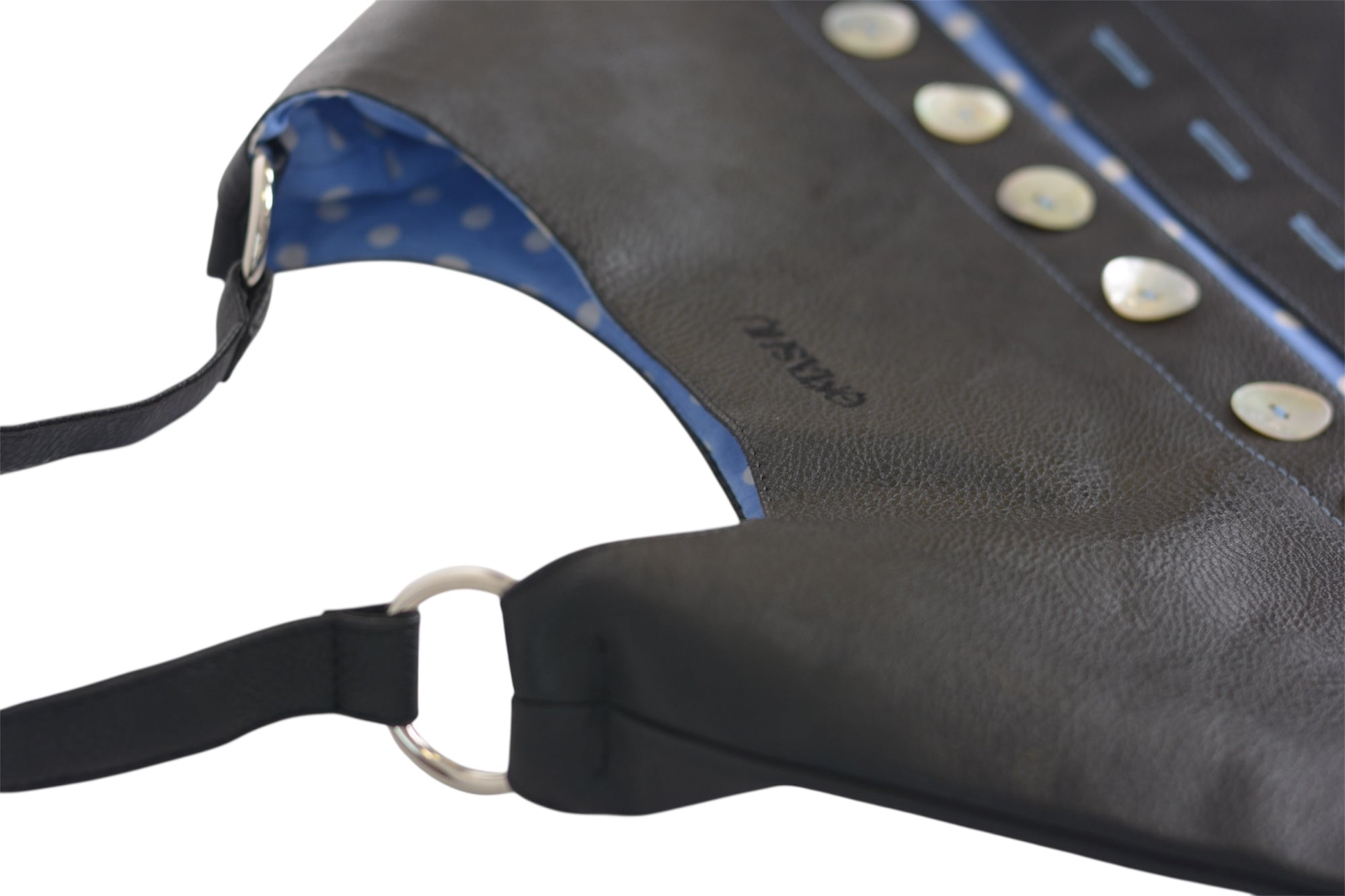 Close-up of Omashu Sylvie bag showing silver O-ring strap attachment, black faux leather, embossed OMASHU logo, and blue polka dot lining edge.