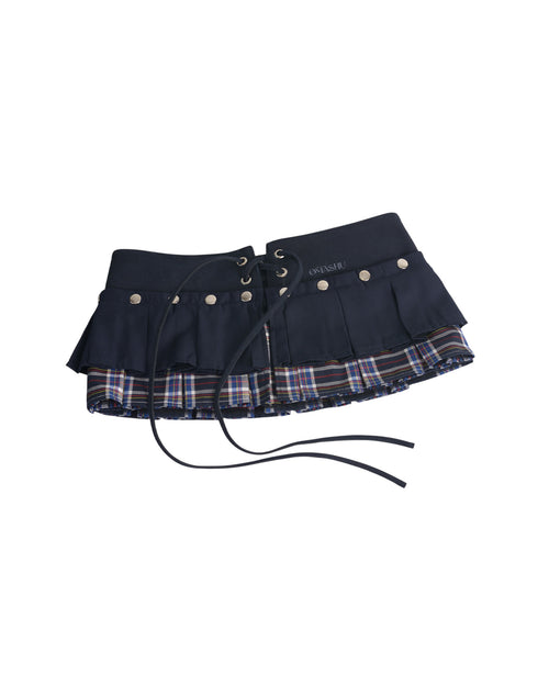 Omashu AYA Belted Layered Skirt, showing a dark navy pleated top mini skirt with silver snap buttons and an integrated corset belt with lacing, layered over a visible red, blue, and white plaid underskirt.
