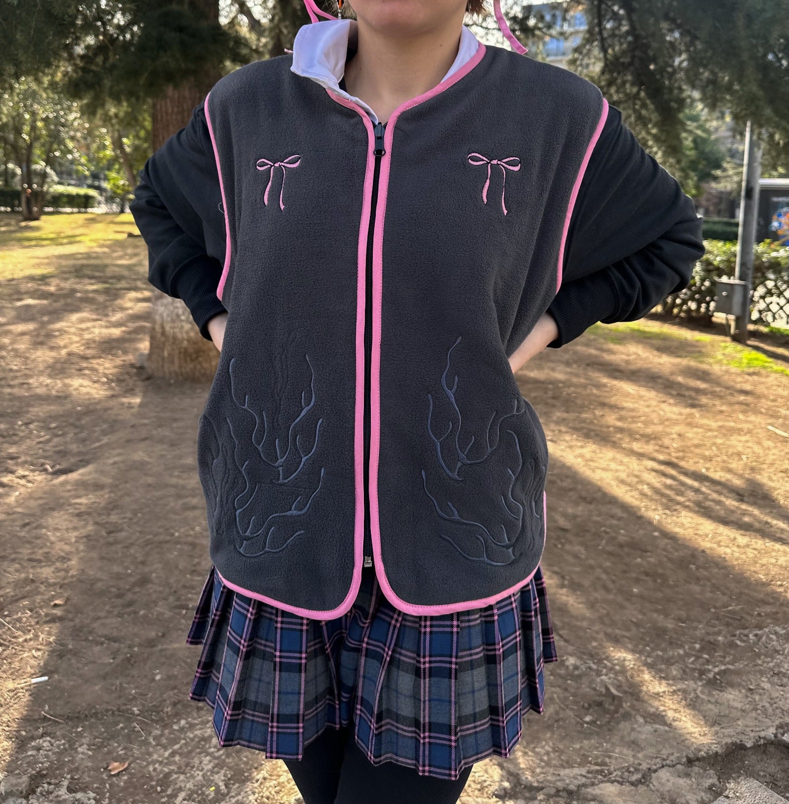 a person wearing a grey jacket with pink outlines and decorative patterns, paired with a plaid skirt, standing outdoors.