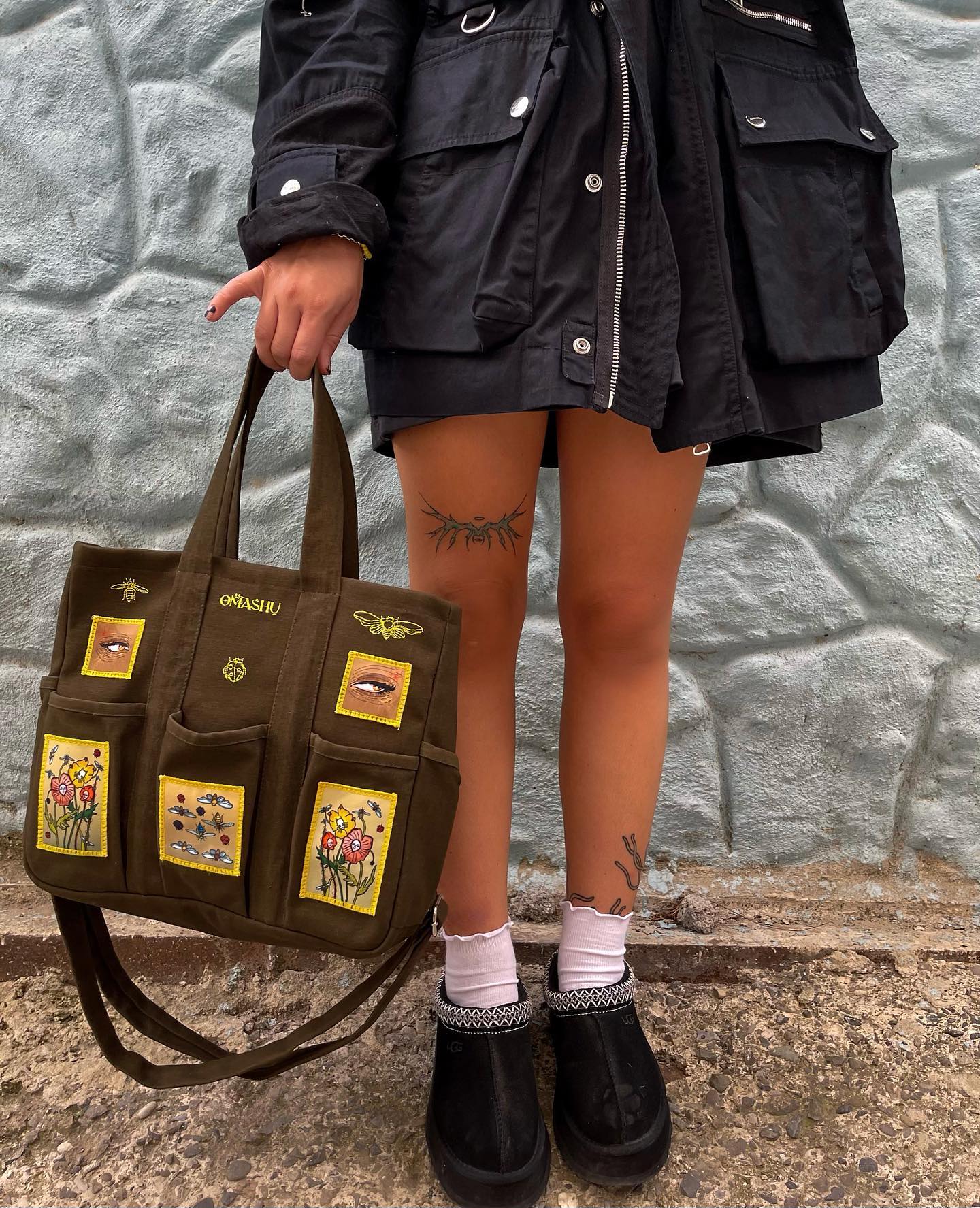 Green tote bag with three patches sewn onto it. The patches are of insects, eyes and a flower. The person holding the bag is wearing a black jacket, black shorts, white socks, and black shoes. The wall in the background is blue and has a rough texture.