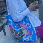 A person wearing a white shirt and pink pants, sitting on a bench and holding a phone in their hand. The person is wearing a blue tote bag with a colorful design of two faces, one pink and one blue, with a bunch of colorful objects in between them. The background consists of a brick wall and a metal railing. tag says 'OMASHU'.