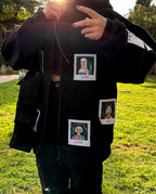 Black hoodie with three square patches on it. The patches are white with a black border and have illustrations of people’s faces and text on them. The top patch has a person with long hair and the text ‘pride’. The middle patch has a person with short hair and the text ‘wrath’. The bottom patch has a person with curly hair and the text ‘sloth’