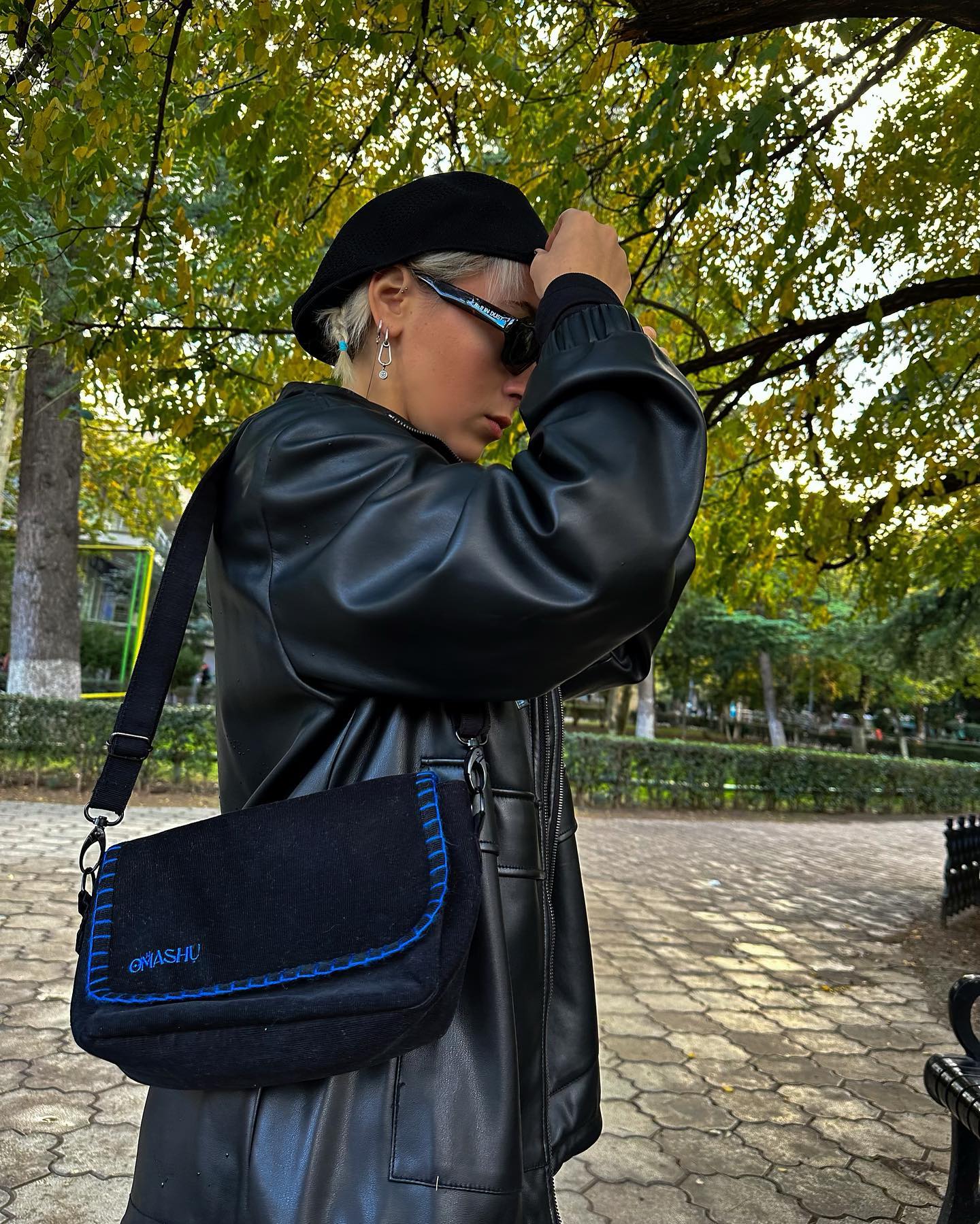 A photo of a person standing in a park with their back to the camera, wearing a black leather jacket, a black hat, and a blue OMASHU crossbody bag with the word ‘OMASHU’ written on it, against a background of trees with green leaves and a park bench.