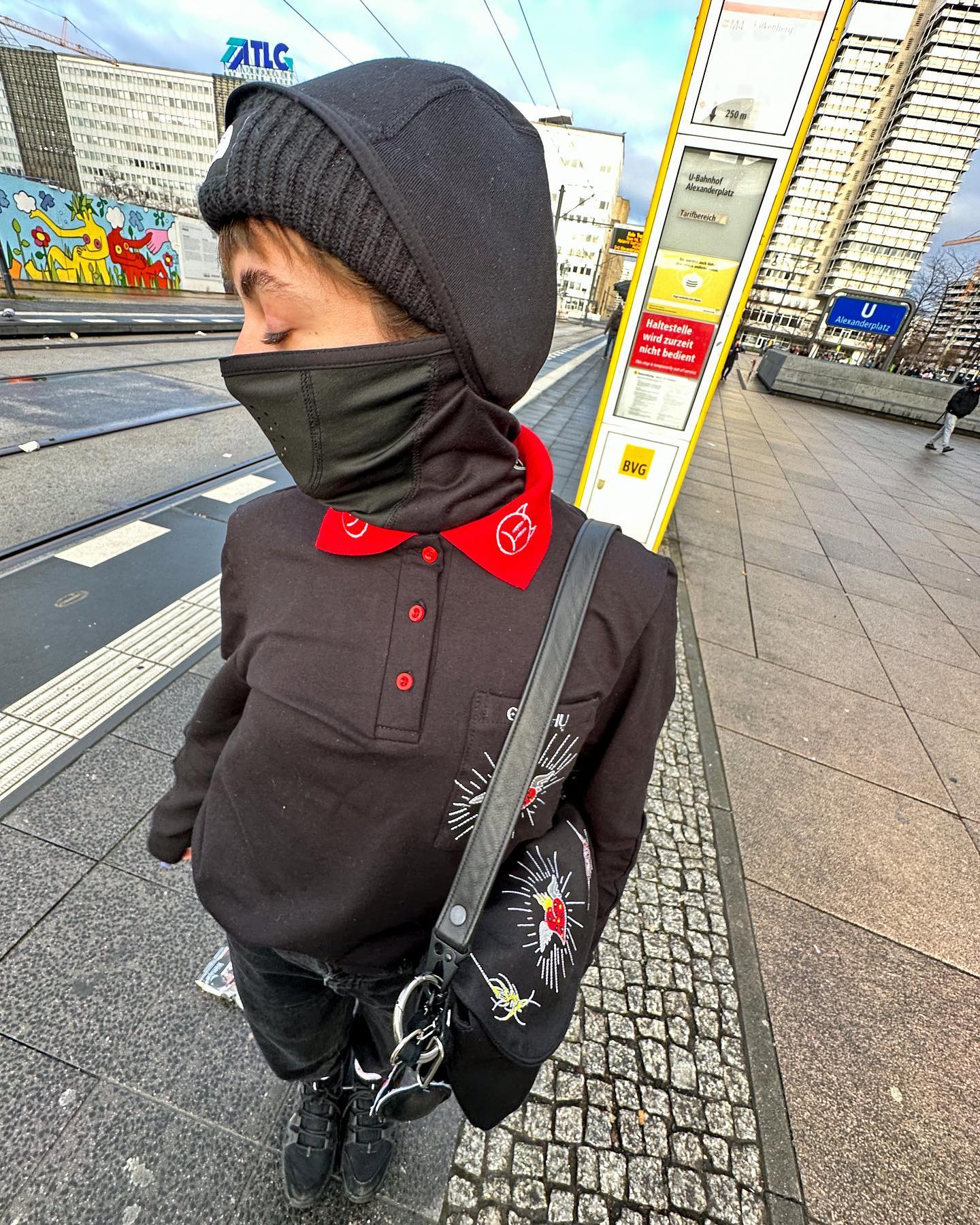 A person wearing a black polo shirt with a red colar. The polo shirt has white designs on it, including a tattooesque figure and a red heart. The person is also wearing black pants and shoes and carrying a black bag with a strap over their shoulder. The background shows a street with buildings and a tram stop. The sky is blue and there are clouds visible.