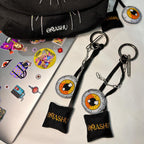 Two keychains on a white surface. The keychains are black and have a rectangular fabric tag with ‘OMASHU’ written on it. The keychains also have a circular pendant with an orange and black eye design. The keychains are attached to a silver metal ring and clip. The background consists of a black bag with white stitching and a laptop covered in colorful stickers.