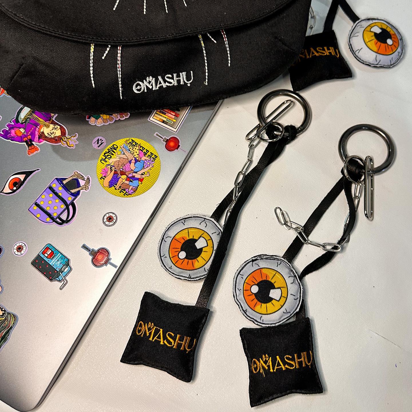 Two keychains on a white surface. The keychains are black and have a rectangular fabric tag with ‘OMASHU’ written on it. The keychains also have a circular pendant with an orange and black eye design. The keychains are attached to a silver metal ring and clip. The background consists of a black bag with white stitching and a laptop covered in colorful stickers.