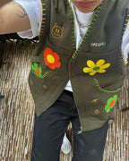 Person wearing green vest with colorful flower designs on it with zipper in front and two pockets on sides, yellow flower on left side and orange flower on right side, small yellow flower on left pocket and small orange flower on right pocket, white shirt, and dark pants, vest has brand name 'OMASHU'