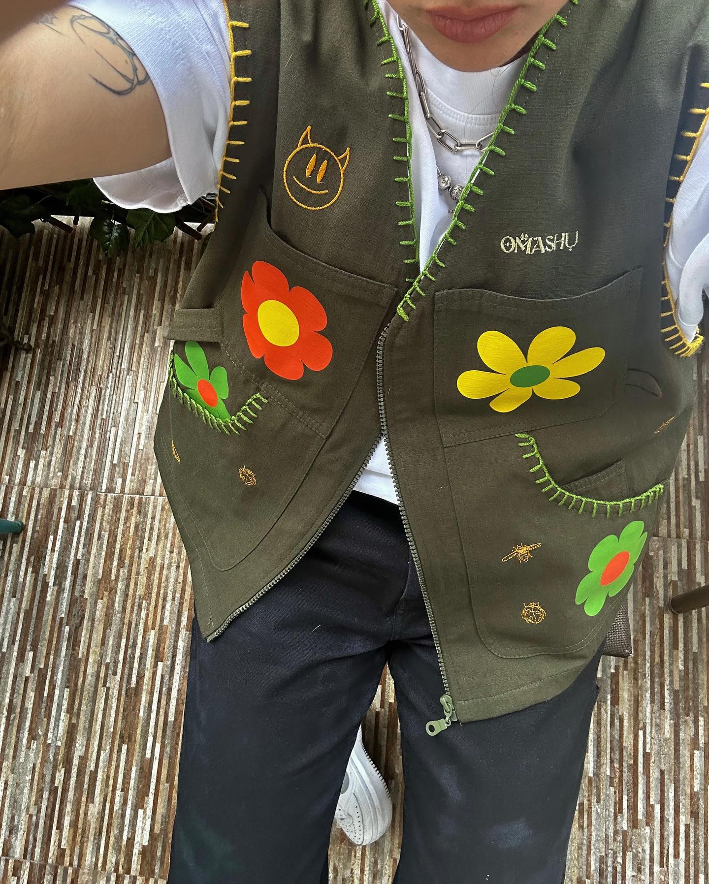 Person wearing green vest with colorful flower designs on it with zipper in front and two pockets on sides, yellow flower on left side and orange flower on right side, small yellow flower on left pocket and small orange flower on right pocket, white shirt, and dark pants, vest has brand name 'OMASHU'