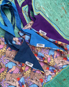 Three colorful tote bags on a turquoise wooden surface. The tote bags have a blue, purple, and orange color scheme. They have a cartoon-like print of people on them. The tote bags have a label that reads ‘OMASHU’ on them.
