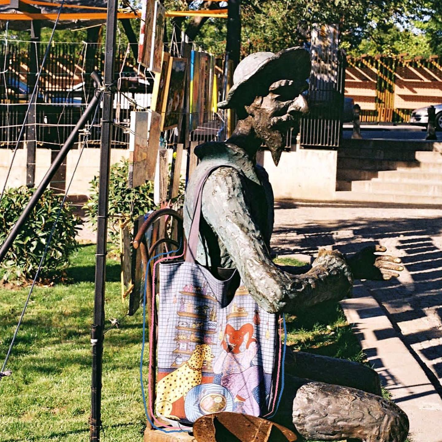 Bronze statue of man with hat and backpack with colorful fox and bird design in park with OMASHU tote bag in background.