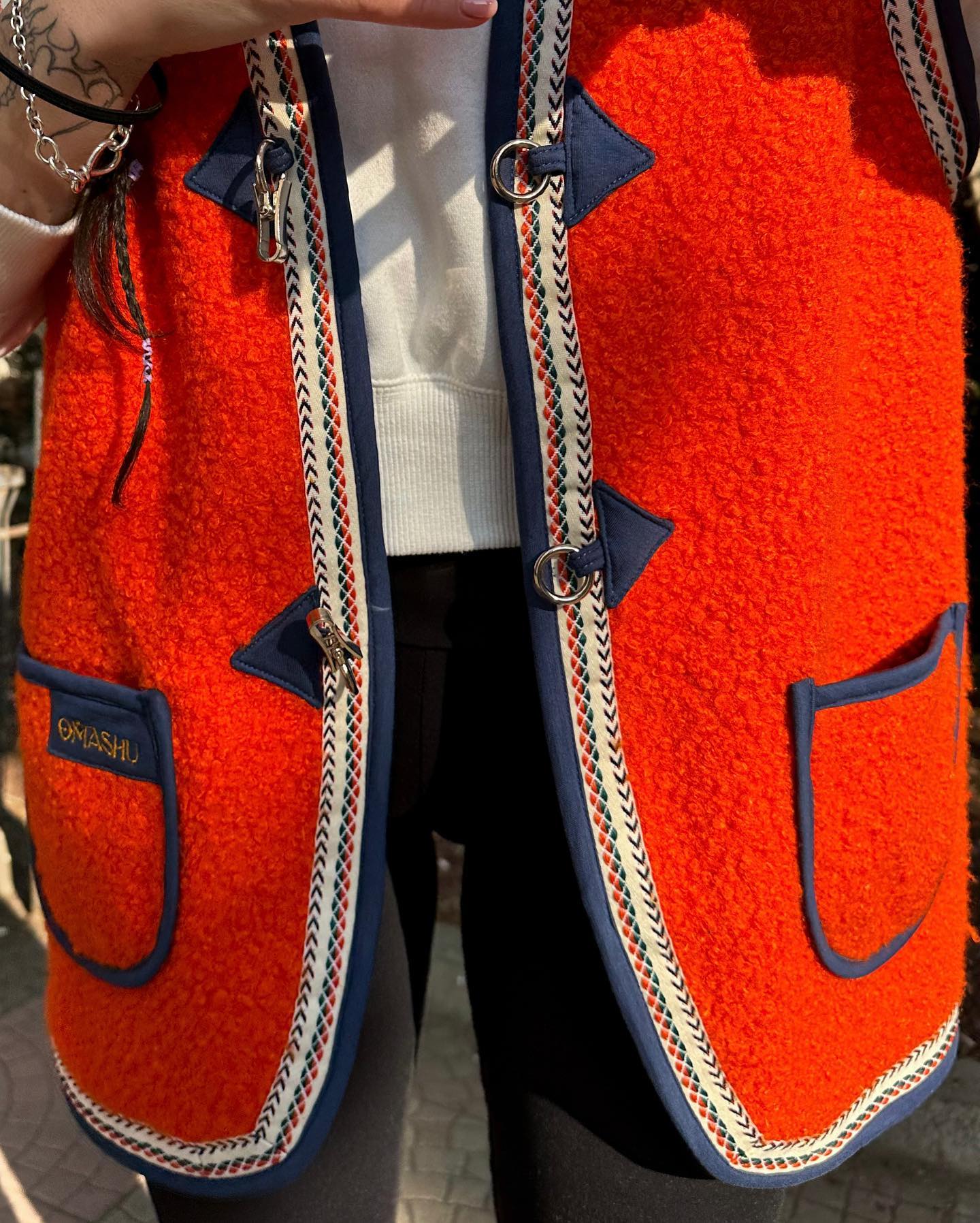 A person wearing a bright orange jacket with blue accents. The jacket has a fuzzy texture and two large pockets on the front. The pockets are outlined in blue and have silver zippers. The jacket also has a blue and white striped trim along the edges. The person is wearing a white shirt underneath the jacket and black pants. The person’s hands are visible, and they are wearing multiple rings and bracelets. The background is a blurred city street.