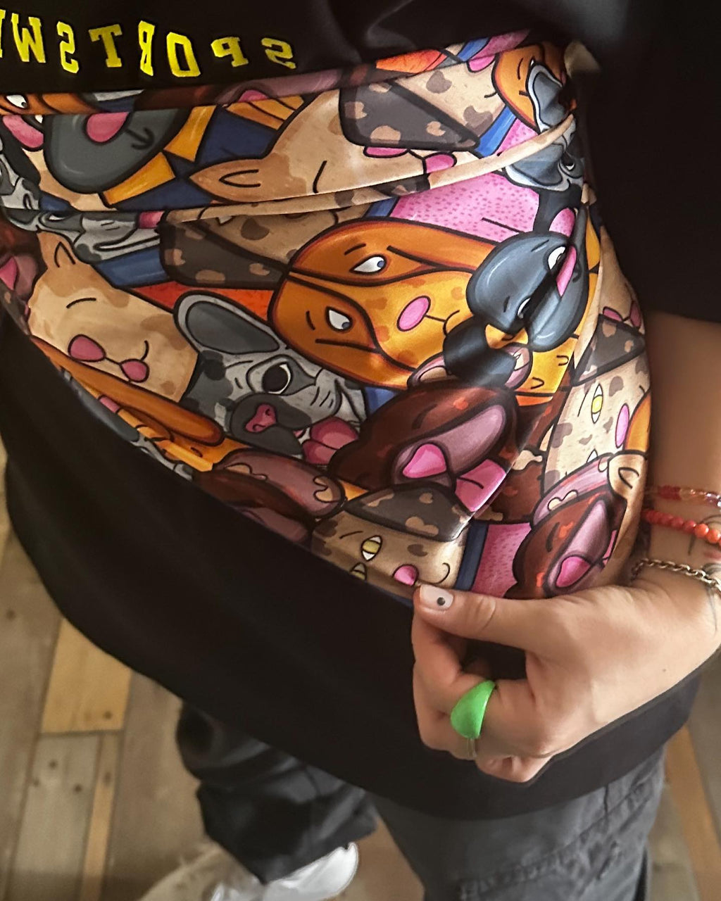 A person wearing a black t-shirt with a colorful cartoon print on it. The print is of various cartoon characters in different poses and expressions. The person is holding the bottom of the t-shirt with their right hand, which has a green ring on the index finger. The background is of a wooden floor and a white wall.