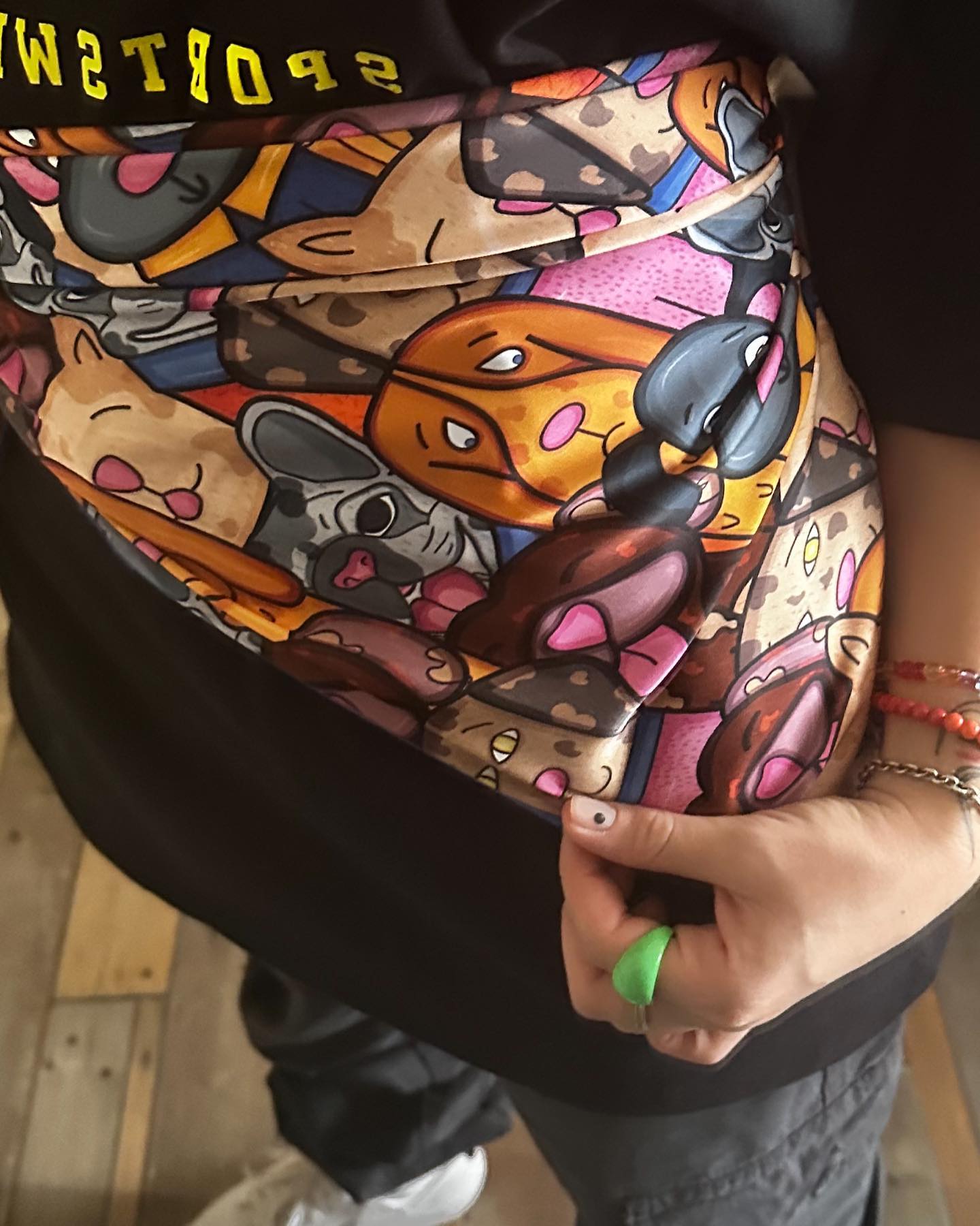 A person wearing a black t-shirt with a colorful cartoon print on it. The print is of various cartoon characters in different poses and expressions. The person is holding the bottom of the t-shirt with their right hand, which has a green ring on the index finger. The background is of a wooden floor and a white wall.
