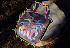 A photo of a colorful tote bag made of brown fabric with a purple handle and green and yellow zigzag borders, featuring a design of a group of people with their arms stretched out in a circle, wearing pink, orange, and yellow clothes, on a tree stump in a forest with fallen leaves, taken during the day with natural lighting.