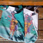 Two tote bags with a colorful illustration on them. The illustration is of a person with a backpack and tentacles coming out of their head. The person is wearing a yellow shirt, blue shorts, and red and white striped socks. The background of the illustration is a light blue color with white bubbles. The tote bags are resting on a wooden bench with a red and green striped cushion. The tote bags have white handles and a white tag on the side that reads ‘Omashu’.