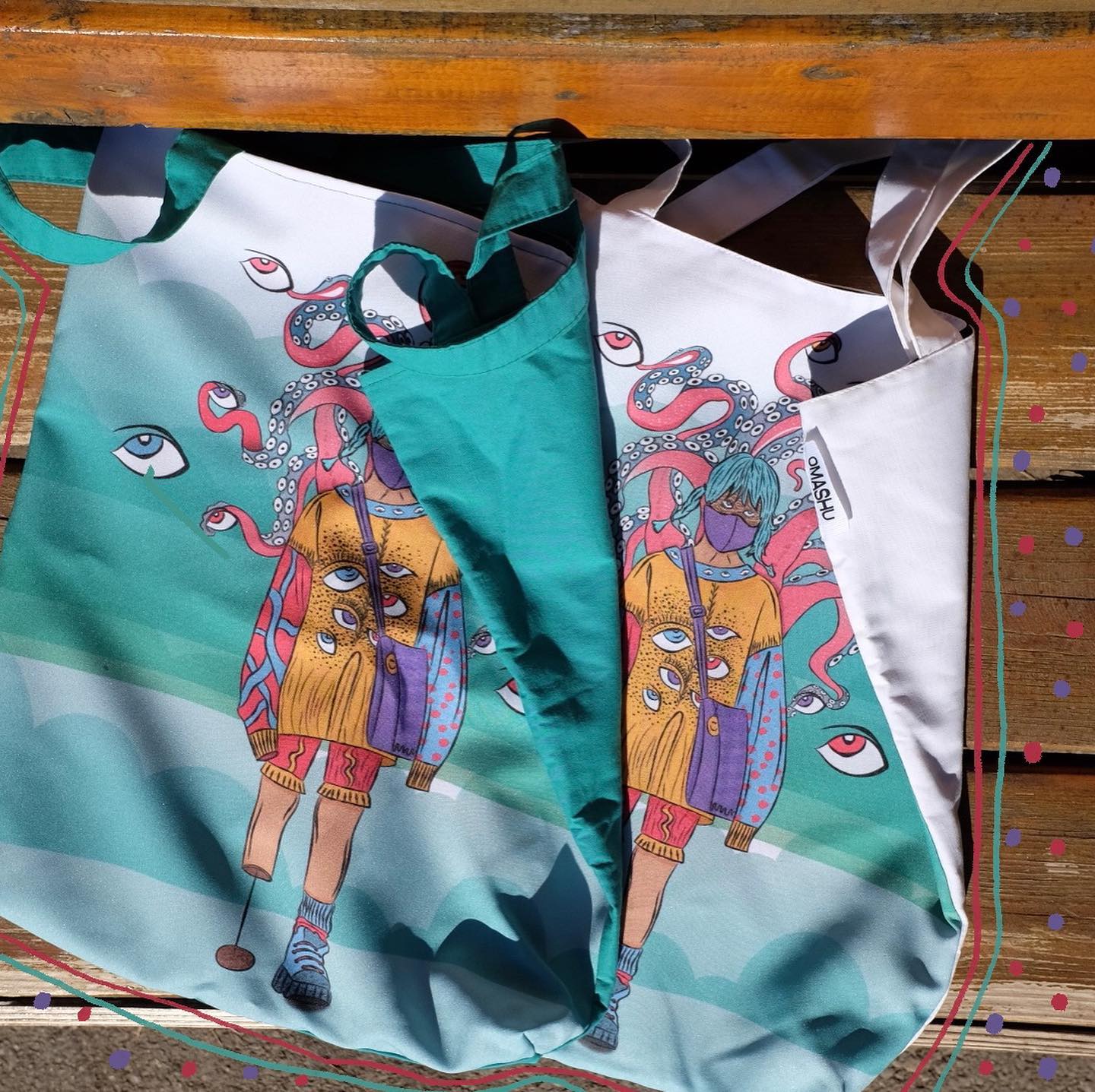 Two tote bags with a colorful illustration on them. The illustration is of a person with a backpack and tentacles coming out of their head. The person is wearing a yellow shirt, blue shorts, and red and white striped socks. The background of the illustration is a light blue color with white bubbles. The tote bags are resting on a wooden bench with a red and green striped cushion. The tote bags have white handles and a white tag on the side that reads ‘Omashu’.
