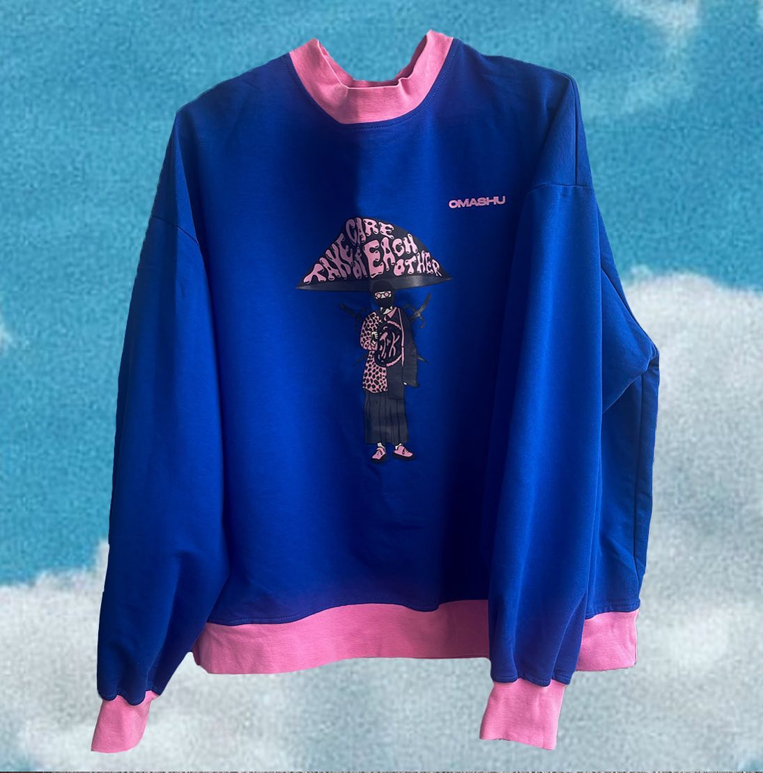 Blue and pink sweatshirt with graphic on front that reads ‘Take Care of Each Other’ in black lettering. Graphic also includes cartoon character with pink hat and skateboard. Small logo on right sleeve that reads ‘OMASHU’. Blue gradient background with cloudy texture.