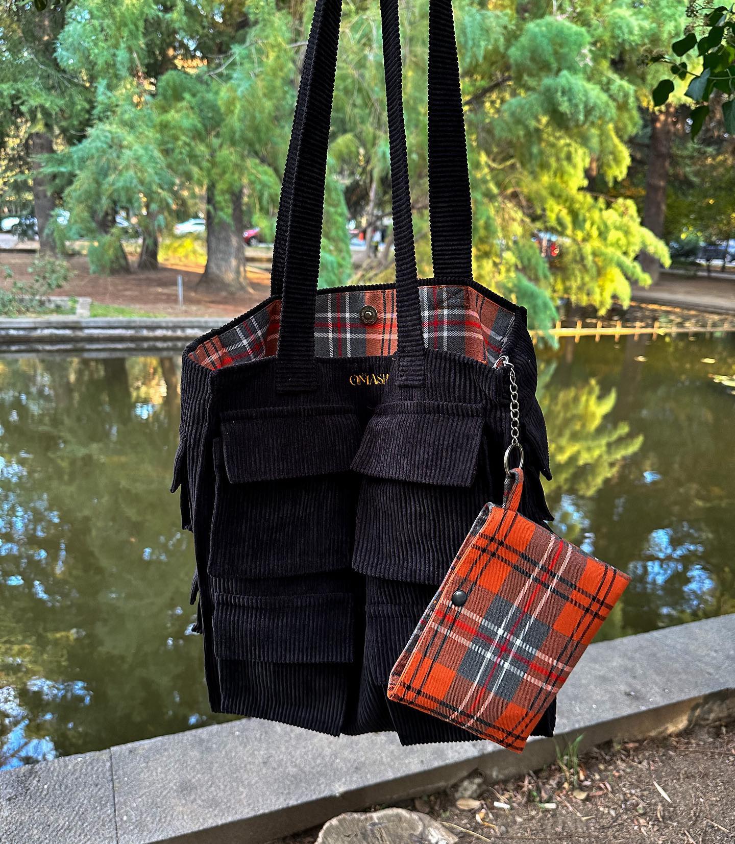 A black and red plaid bag with a matching wallet on a stone wall in a park setting. The bag has two black straps and a black flap with a gold button. The wallet is red plaid with a gold chain. The background consists of a pond with trees and bushes. The stone wall is gray and has a rough texture.