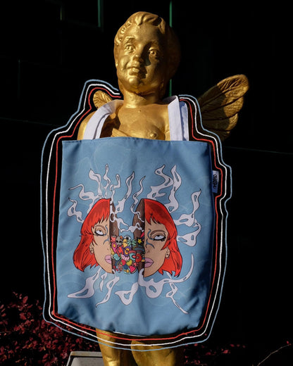 A blue tote bag with a graphic design of a red-haired woman with flowers in her hair. The woman’s face is blurred out to protect her privacy. The tote bag is hanging on a golden statue of a person with wings. The background is a dark green wall with red leaves scattered on the ground.