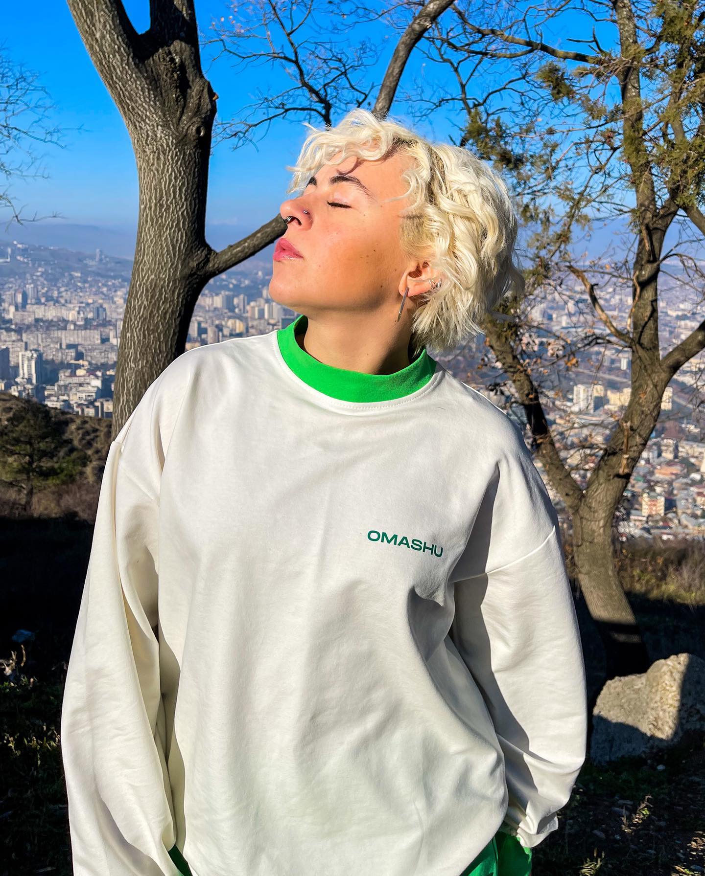 A person standing in front of a tree with a cityscape in the background. The person is wearing a white sweatshirt with green text that reads ‘OMASHU’ and green sweatpants