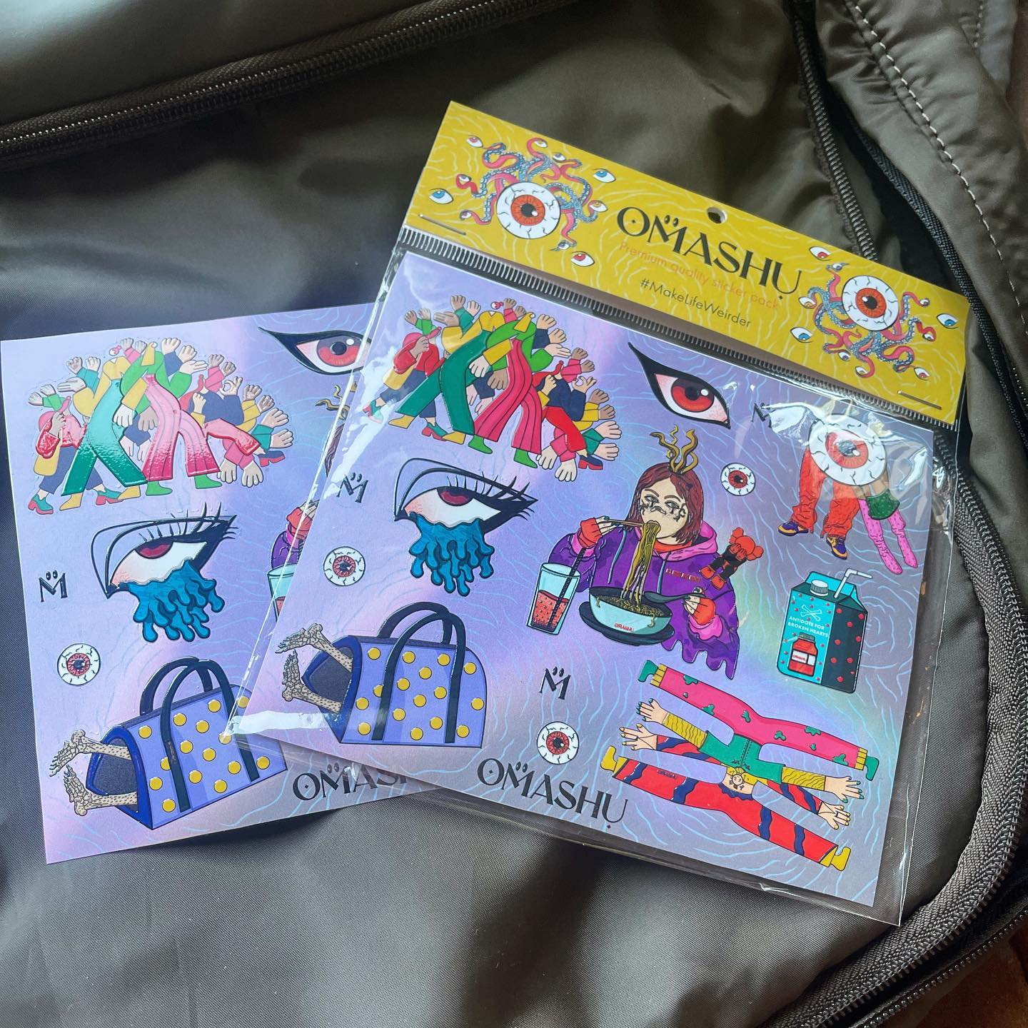 A photo of two colorful sticker packs on a black background. The postcards are illustrated with vibrant colors and patterns depicting a pair of eyes, a handbag, and a pair of shoes. The sticker pack is in a clear plastic packaging with a yellow label that reads ‘OMASHU’. The stickers inside are also illustrated with vibrant colors and patterns. The background is a black fabric, possibly a bag or a jacket. You can see the zipper and stitching details.