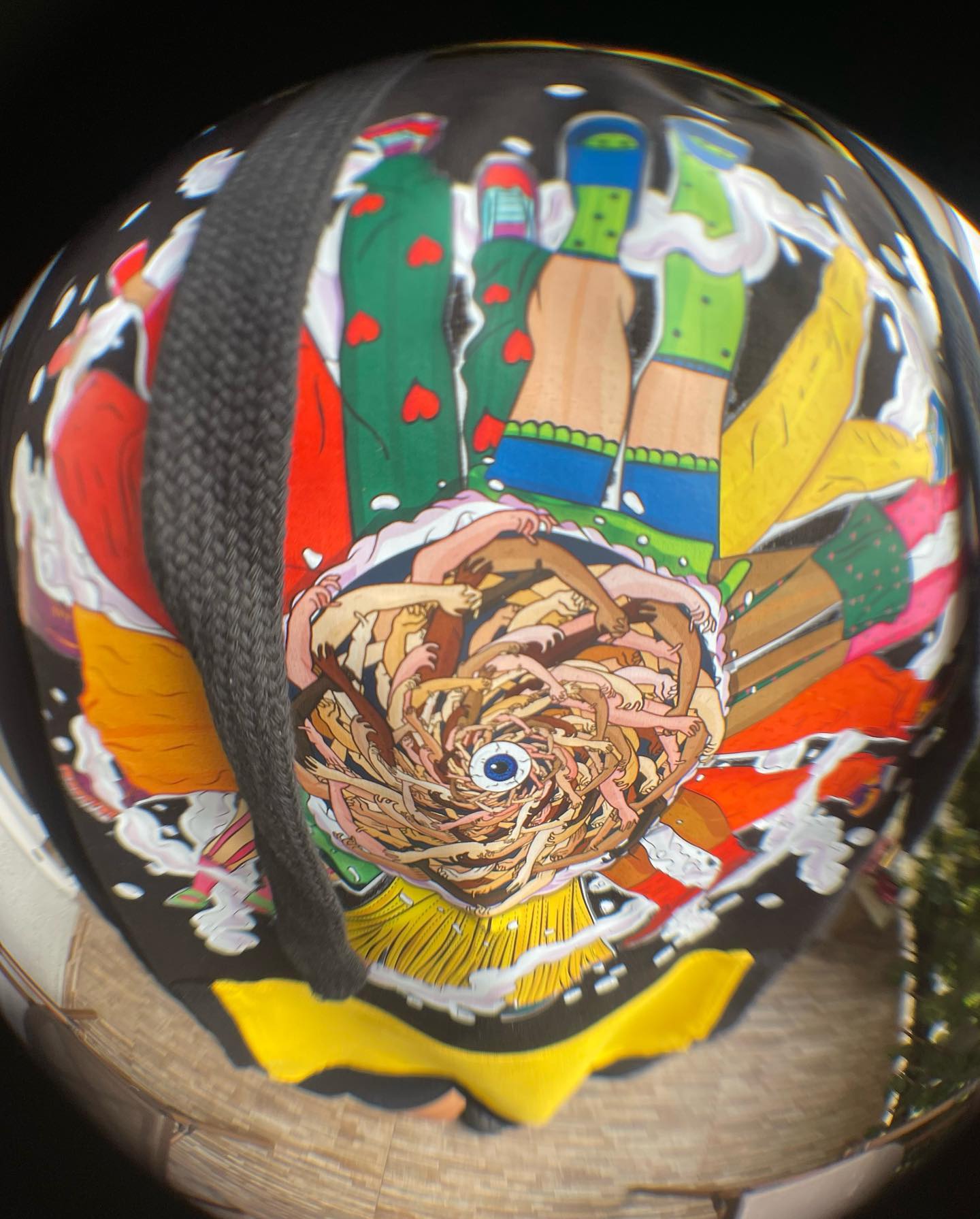 A colorful helmet with a fish eye lens effect. The helmet is black with a colorful design on it. The design features a large rose in the center with an eye in the middle. The design also features a rainbow, a castle, and a green snake. The helmet is sitting on a wooden surface.