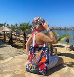 A person sitting on a rocky beach with palm trees and beach umbrellas in the background. The person is wearing a white dress and a colorful headscarf. The person is holding a large tote bag with a colorful design on it. The bag has a large image of a snail on it, surrounded by flowers and leaves. The person is looking out towards the ocean, with their hand shielding their eyes from the sun. The sky is clear and blue, and the water is calm.