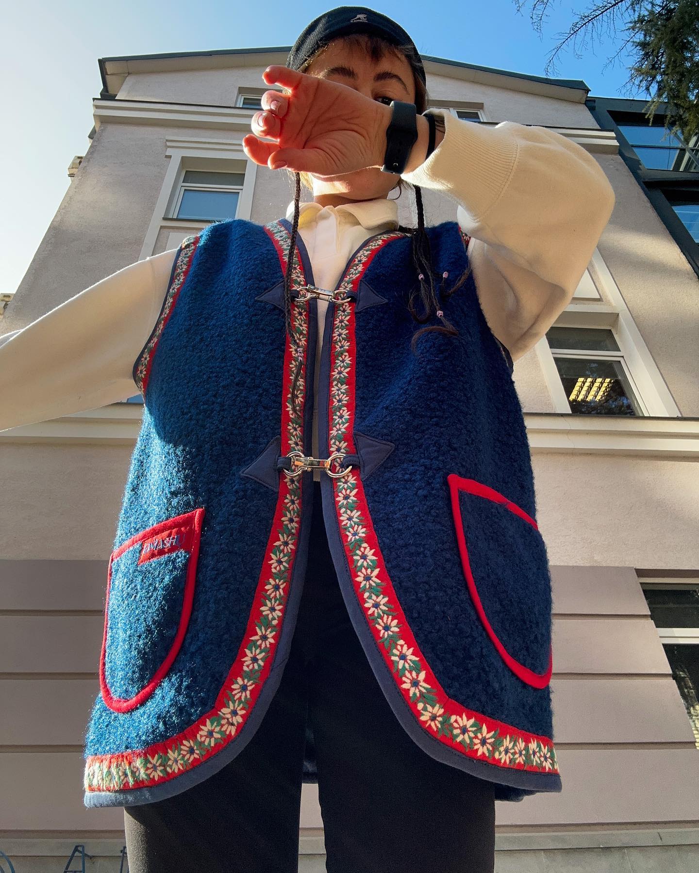 A person wearing a blue vest with red trim and a white shirt. The vest has a red pocket on the left side and a red and white floral pattern on the bottom. The person is holding a brown rectangular object in front of their face, obscuring it. The background is a building with a white facade and a blue sky.
