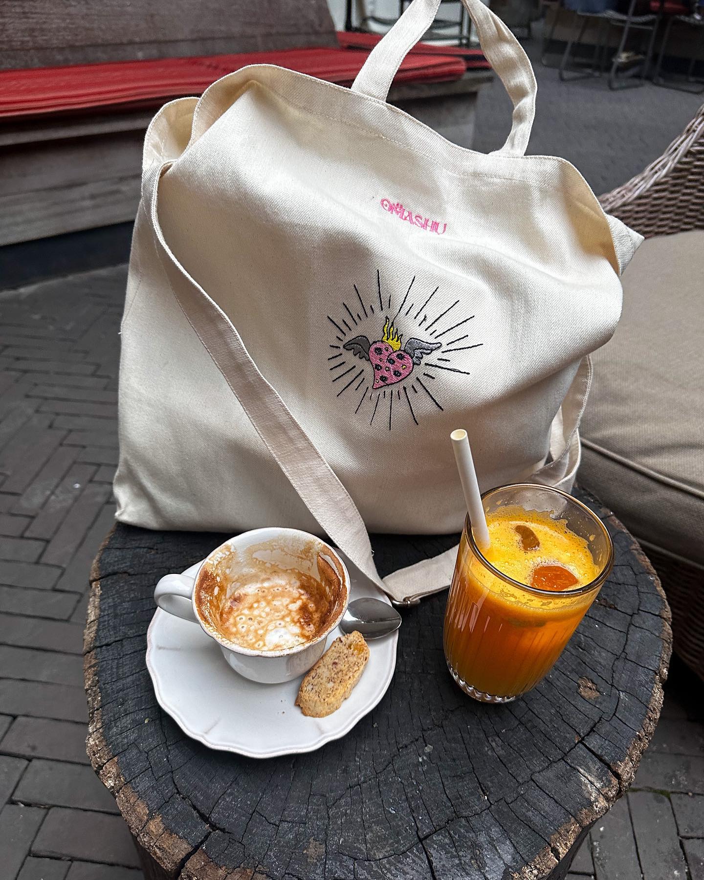 A table made of a tree trunk with a beige OMASHU tote bag featuring a pink and black logo on top, a white cup of coffee with a handle and spoon, half empty with a biscuit on the saucer, and a tall glass of orange juice with ice cubes and a slice of orange.