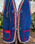 A person wearing a blue vest with red and white floral trim. The vest is made of a blue textured fabric and has two red pockets with white floral trim. It is fastened with two silver clasps at the front. The person is also wearing a white shirt and black pants. The background is a garden with trees and bushes.
