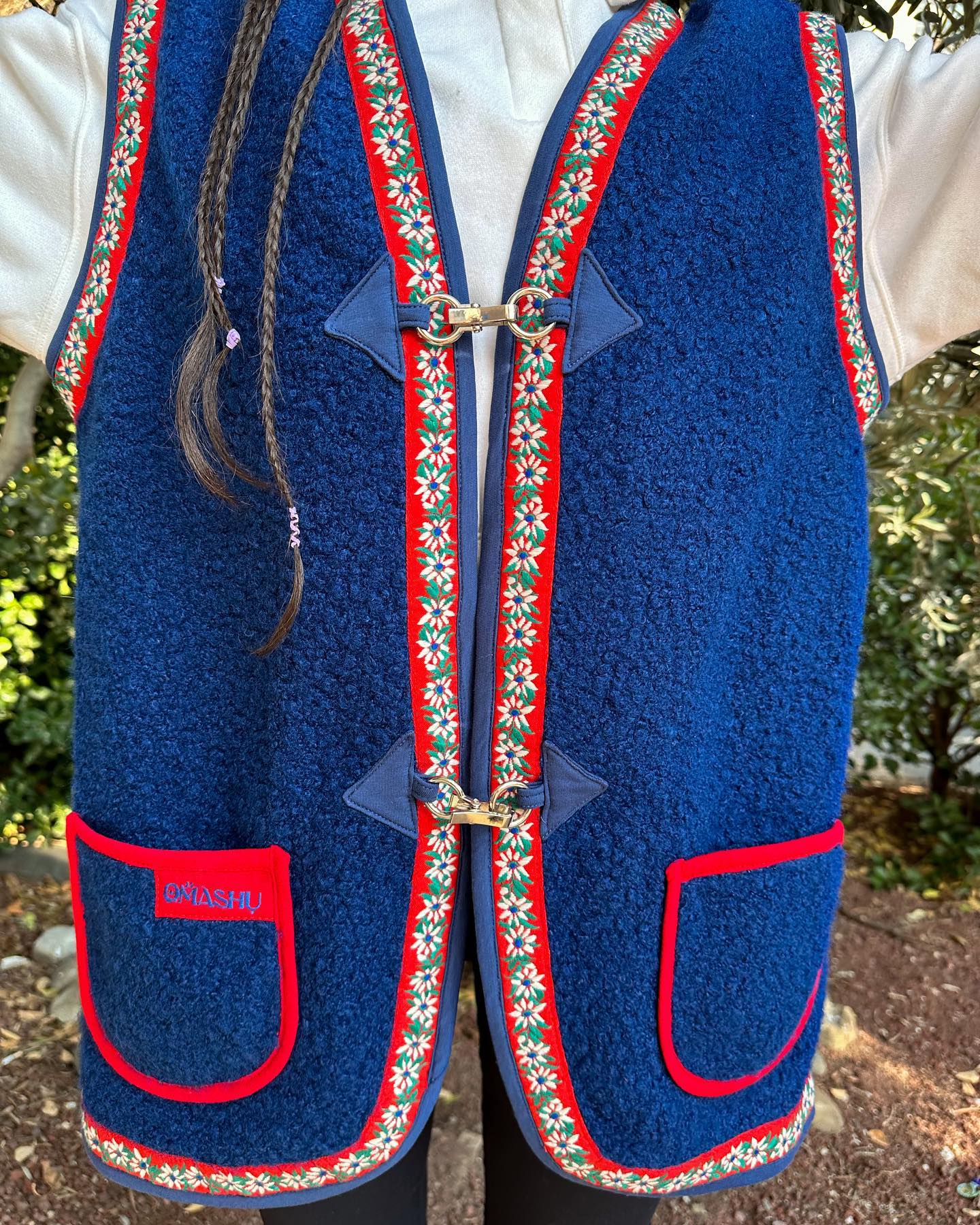 A person wearing a blue vest with red and white floral trim. The vest is made of a blue textured fabric and has two red pockets with white floral trim. It is fastened with two silver clasps at the front. The person is also wearing a white shirt and black pants. The background is a garden with trees and bushes.