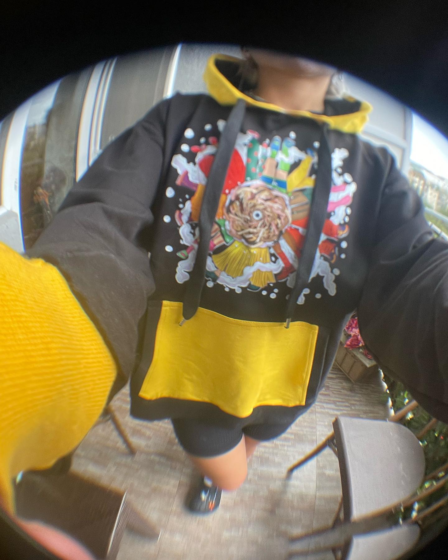 A person wearing a black hoodie with a colorful graphic on it. The graphic is of a lion’s head with a crown and red and white stripes behind it. The person is also wearing a yellow skirt and black shoes. The photo is taken with a fisheye lens, distorting the edges of the image. The background is a patio with plants and furniture.