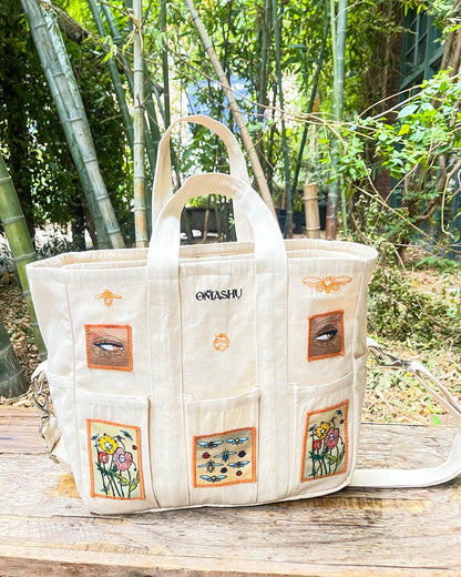 OMASHU white carry-all tote bag with colorful patches sewn onto it. The patches include two orange flowers, a blue fish, and a pink flower. The tote bag has two handles and a shoulder strap. The background is a bamboo grove with green leaves and brown stems. The tote bag is resting on a wooden bench with a chain attached to it.