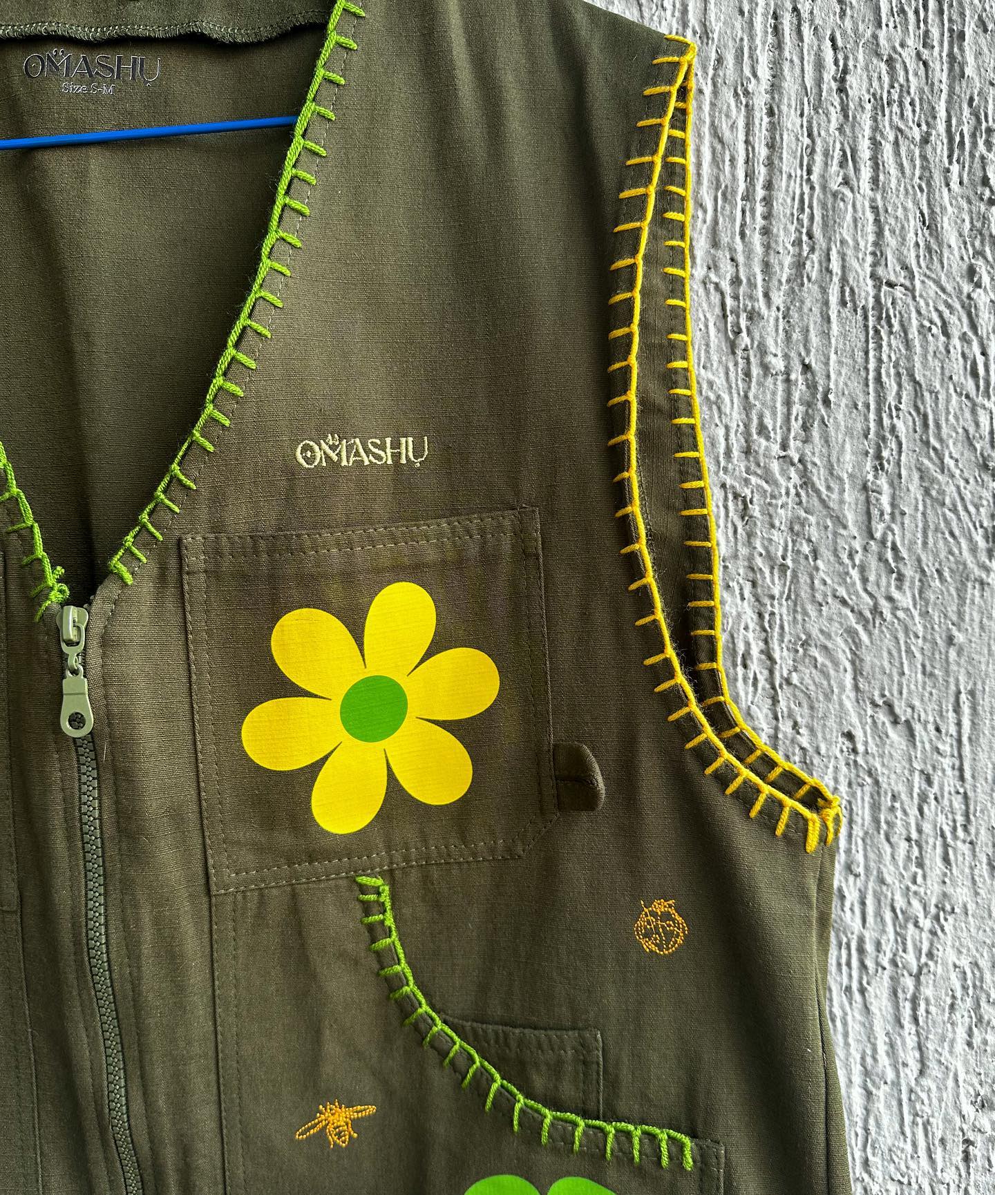 Green vest with yellow stitching and yellow flower on pocket with zipper on front and small logo on bottom right corner that reads ‘OMASHU’ hanging on blue hanger.