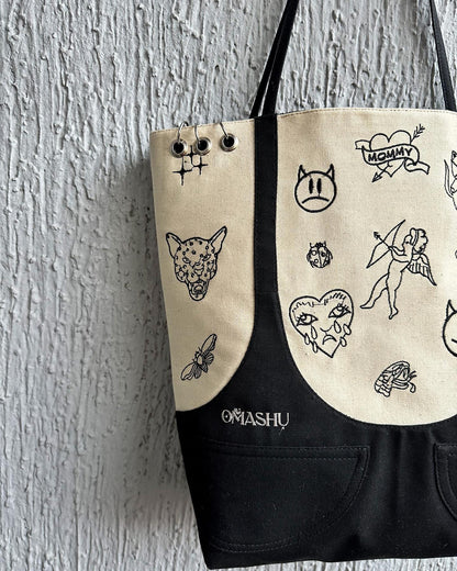 A canvas tote bag with a black base and straps. The bag has various doodles and illustrations on it, including a cat, a butterfly, and a fairy. The illustrations are in black ink and are scattered across the bag. The bag has a label on the bottom right corner that reads ‘OMASHU’. The bag has three silver safety pins attached to the top left corner.