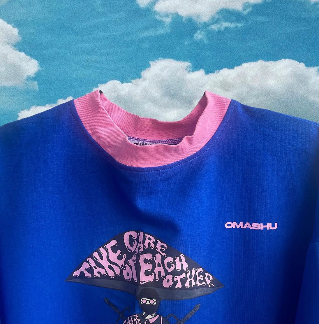 Blue t-shirt with pink collar and graphic of cartoon character with black and white striped shirt and sunglasses. Character is holding a sign that reads ‘Take care of each other’. Small logo on right side that reads ‘OMASHU’. Blue sky background with white clouds.