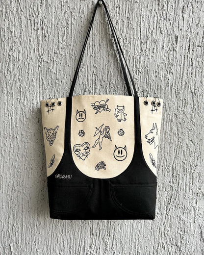 A canvas tote bag with a black bottom and straps. The top part of the tote bag is beige and has various doodles of animals and objects on it. The doodles include a cat, a dog, a fish, a bird, a flower, a mushroom, a butterfly, and a smiley face. The tote bag has a label on the bottom right corner that reads ‘OMASHU’.”