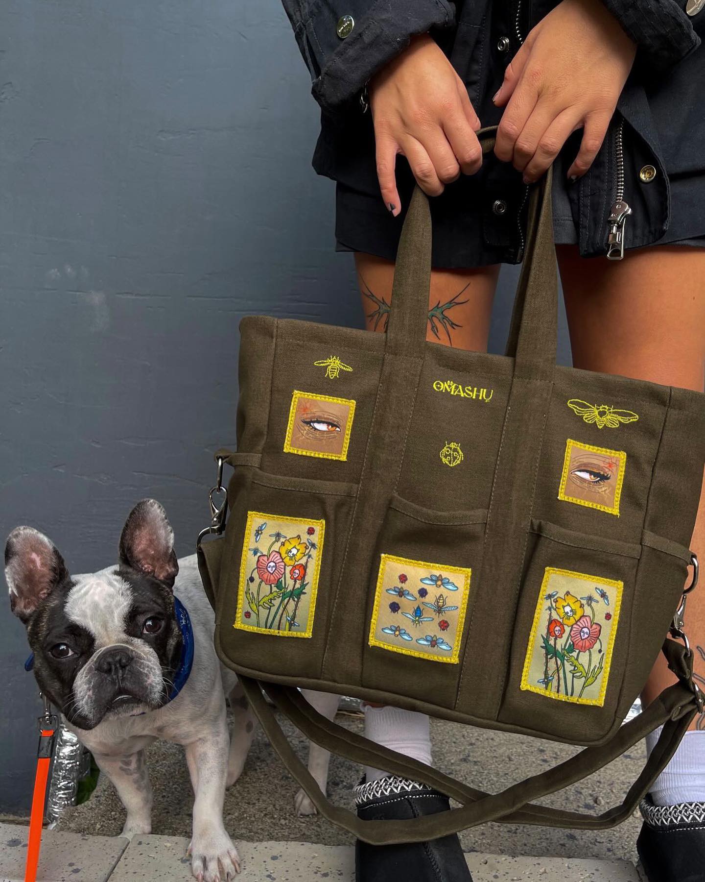 Green tote bag with four square patches on it with colorful flower designs. The patches are yellow, pink, and blue in color. The bag has a label on it that reads ‘OMASHU’. The person holding the bag is standing next to a black and white French bulldog wearing a blue collar. The background is a gray wall with a black railing.