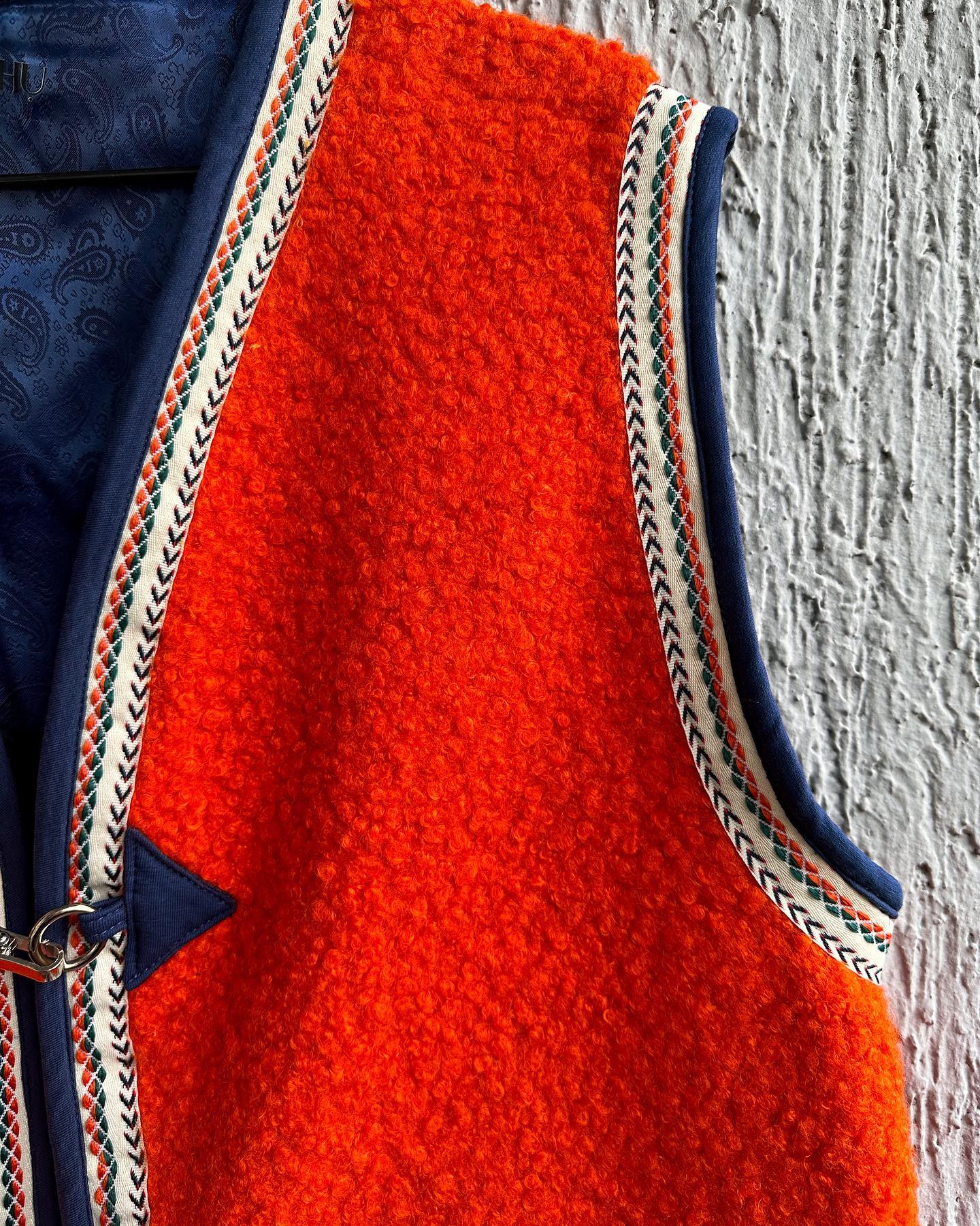 Close-up of an orange felt vest with a blue and white braided trim. The vest has a blue strap with a silver buckle. The background is a white textured wall. The image is taken from the side, so only part of the vest is visible.