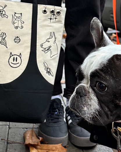 A black and white French Bulldog on a leash, standing on a brick sidewalk. The dog is looking up at the camera with its head tilted to the side. The dog has a black collar with a silver tag. The dog is standing next to a person’s legs, who is wearing black pants and black sneakers. The person is carrying a beige tote bag with black illustrations of cats, dogs, and bees on it. There is a fallen leaf on the ground next to the dog’s paw.