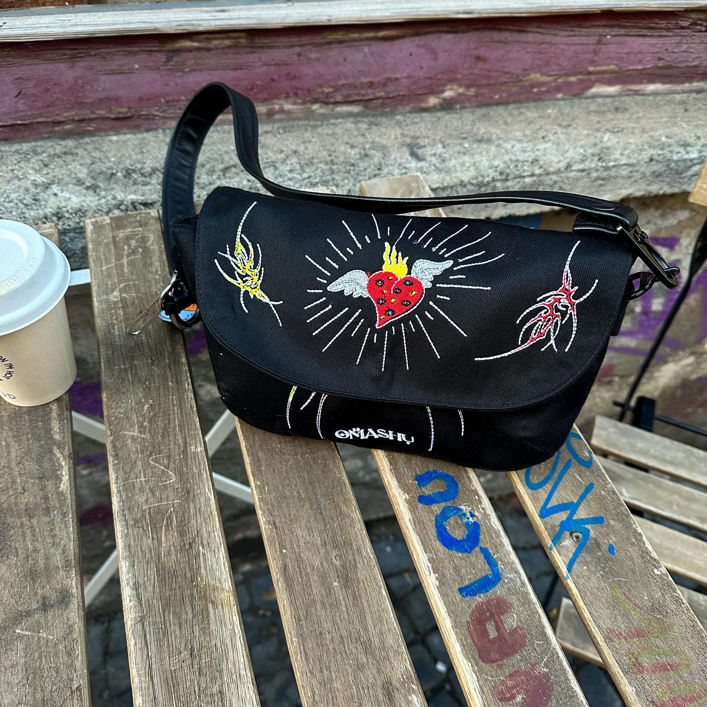 A black OMASHU crossbody bag with colorful designs including a yellow figure, a red figure, and a red heart, rests on a weathered wooden bench with a coffee cup next to it against a brick wall background.